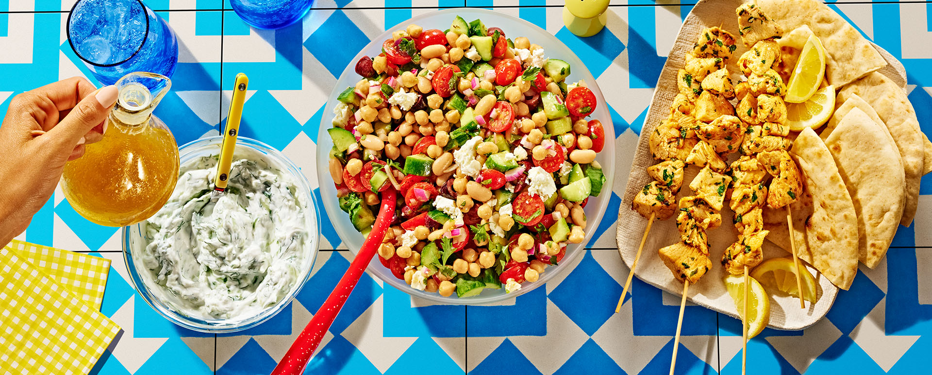 Dense bean salad next to a bowl of tzatziki sauce and a plate of chicken skewers.